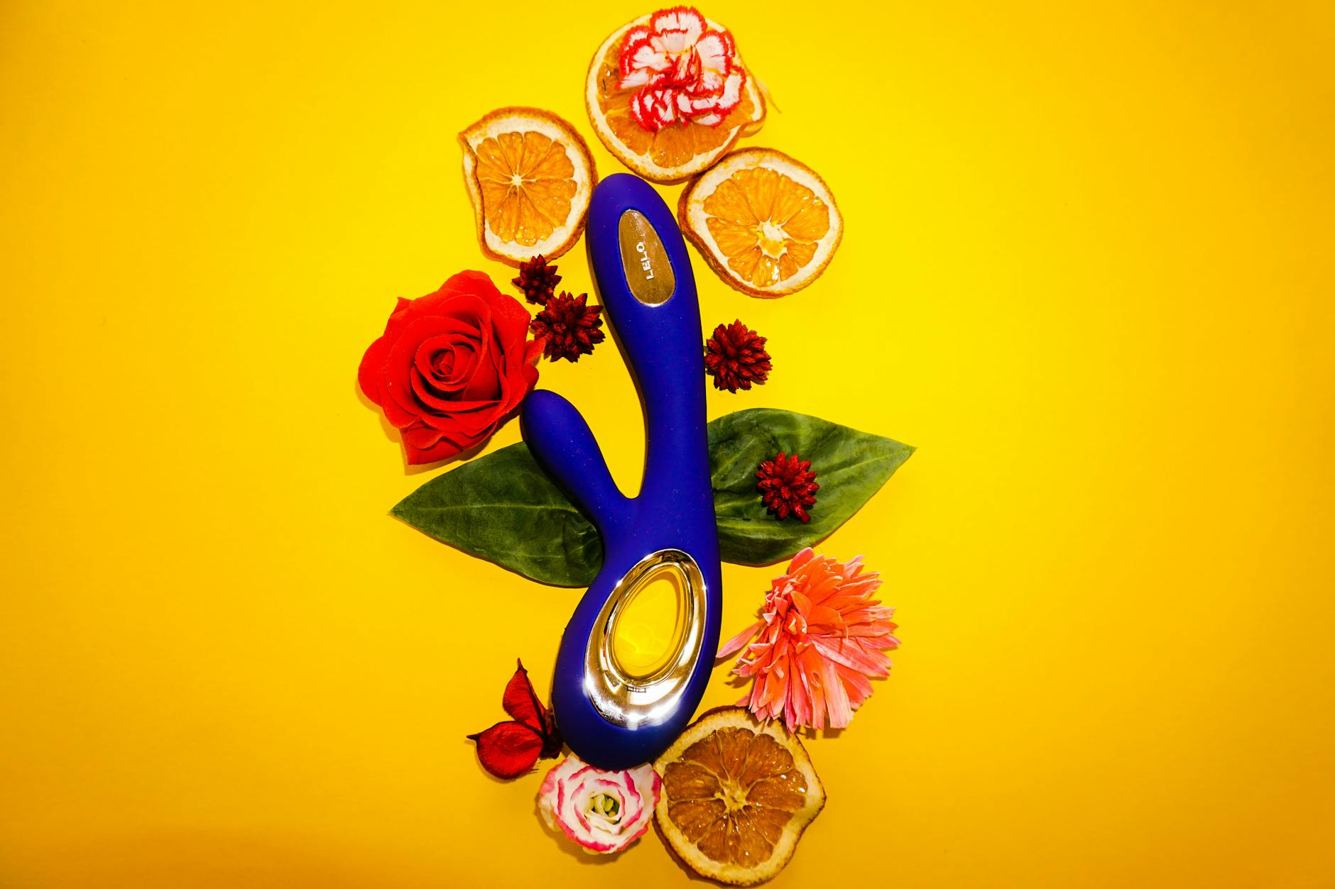 Un vibratore blu circondato da fiori e fette di arancia su uno sfondo giallo