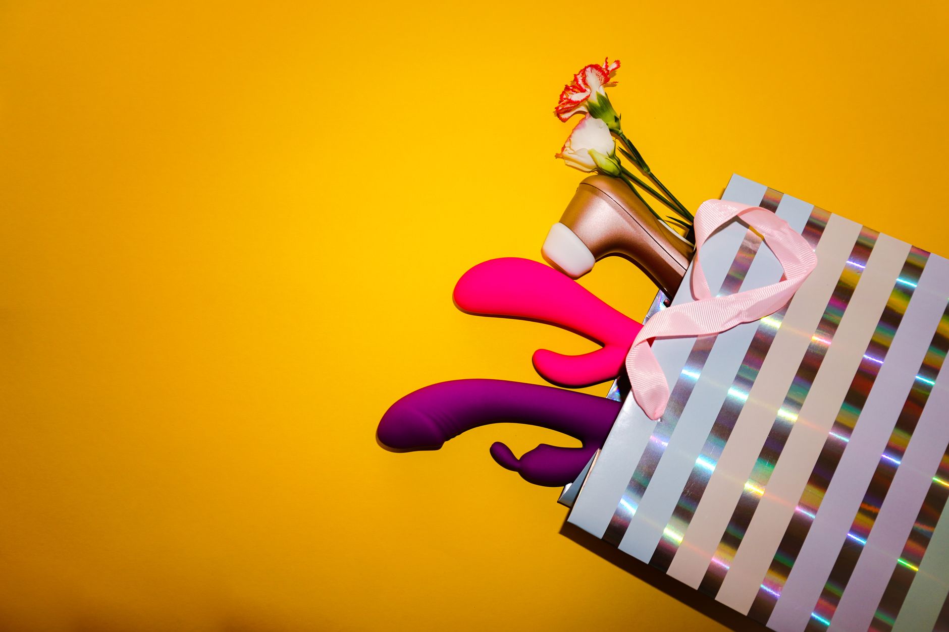 Colorful clitoral vibrators with flowers in a holographic gift bag against a bold yellow background